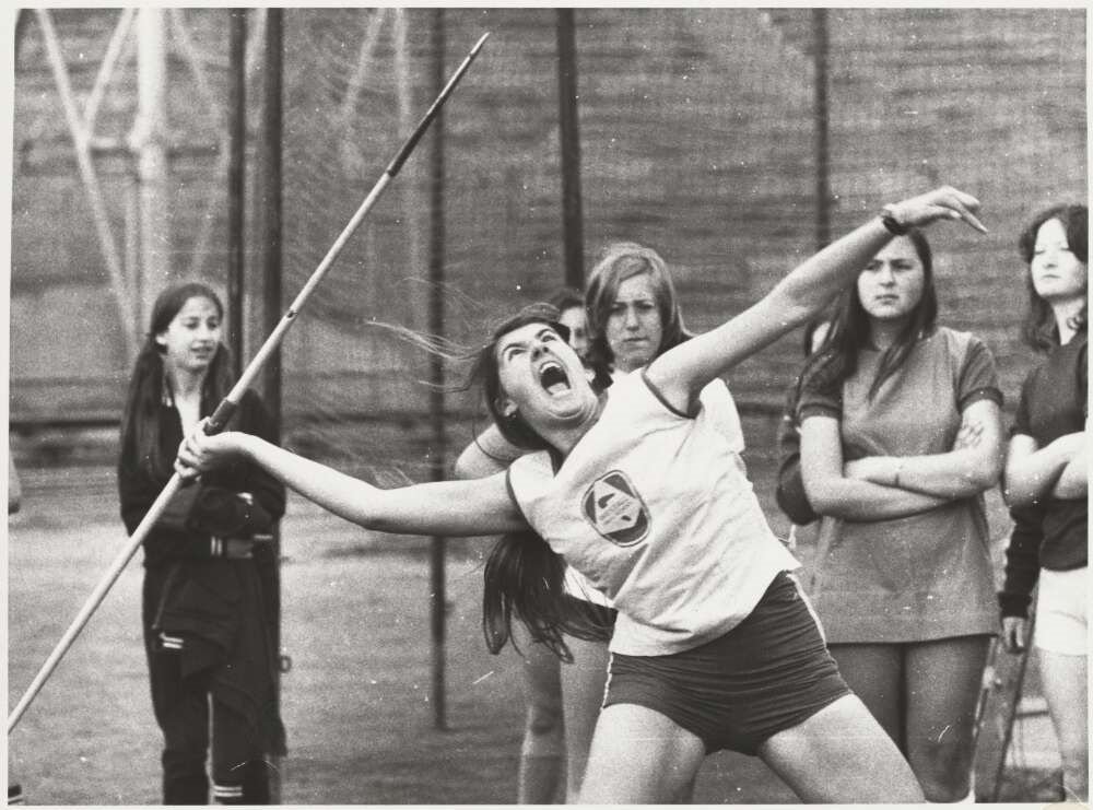 A black and white photo of a girl leaning back to throw a javelin while yelling.