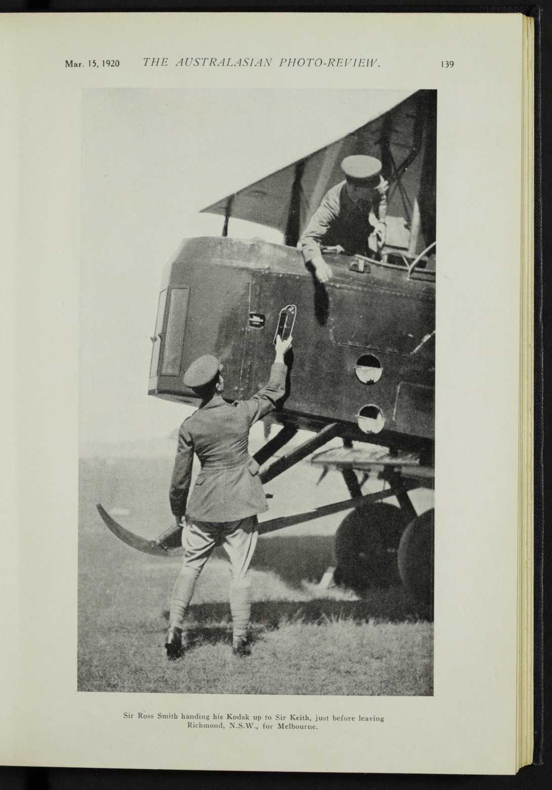 Man in military uniform standing next to front of plane, reaching up with camera in his hand while man in cockpit reaches down to grab it