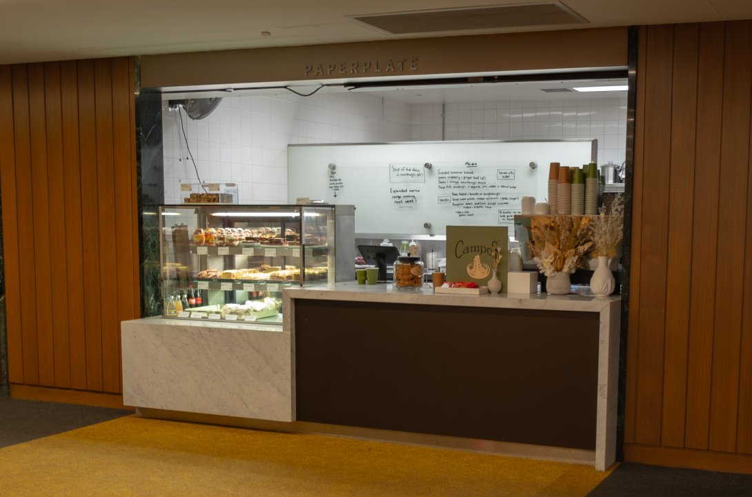 Cafe counter with a display of cakes, slices and fruit salad as well as a coffee machine, stacks of cups and flowers on the counter