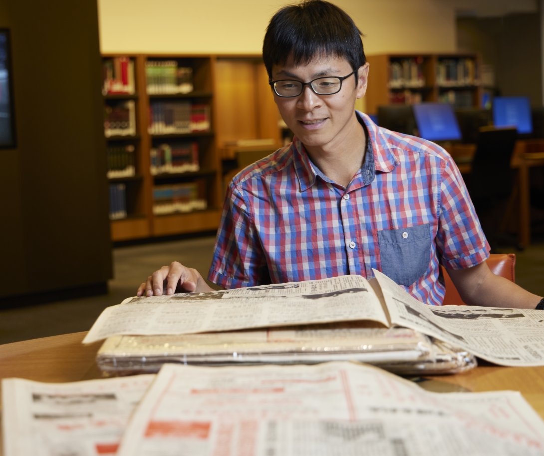 Man with short black hair and black glasses in a short-sleeved checkered shirt sitting at a desk and reading a Chinese language newspaper