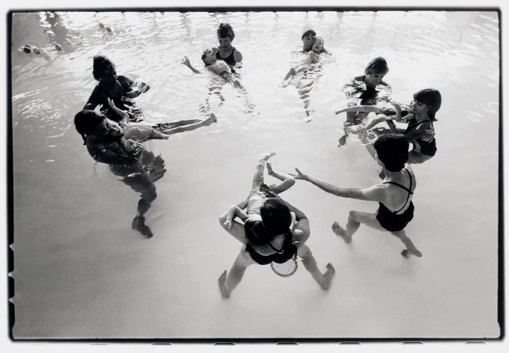 A black and white landscape of women floating in a pool in a circle with toddlers accompanying them.