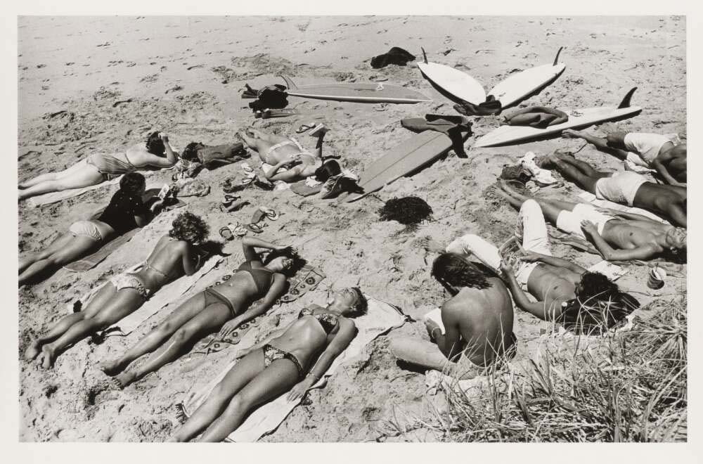 A black and white landscape of a group of sunbathing people lying on a beach with multiple surfboards lying on the ground together.