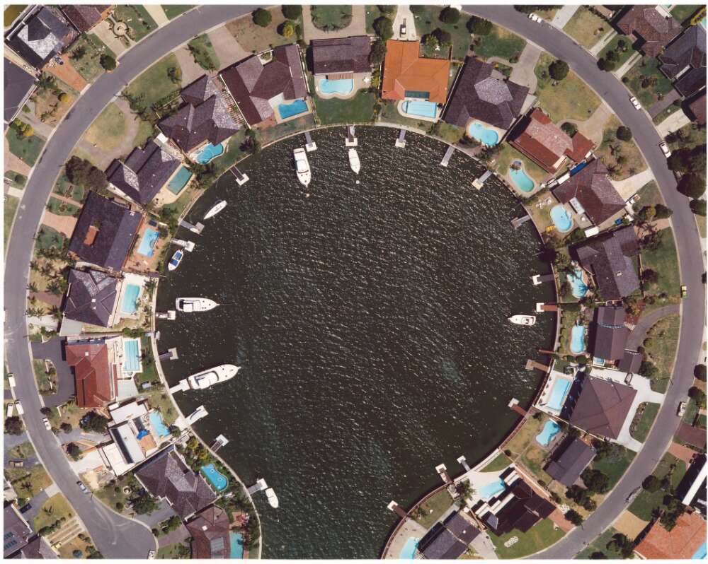 An aerial shot of a suburb. The houses sit in a circle around a round body of water with an opening for the water at the base of the image. there are boats at piers in the water.