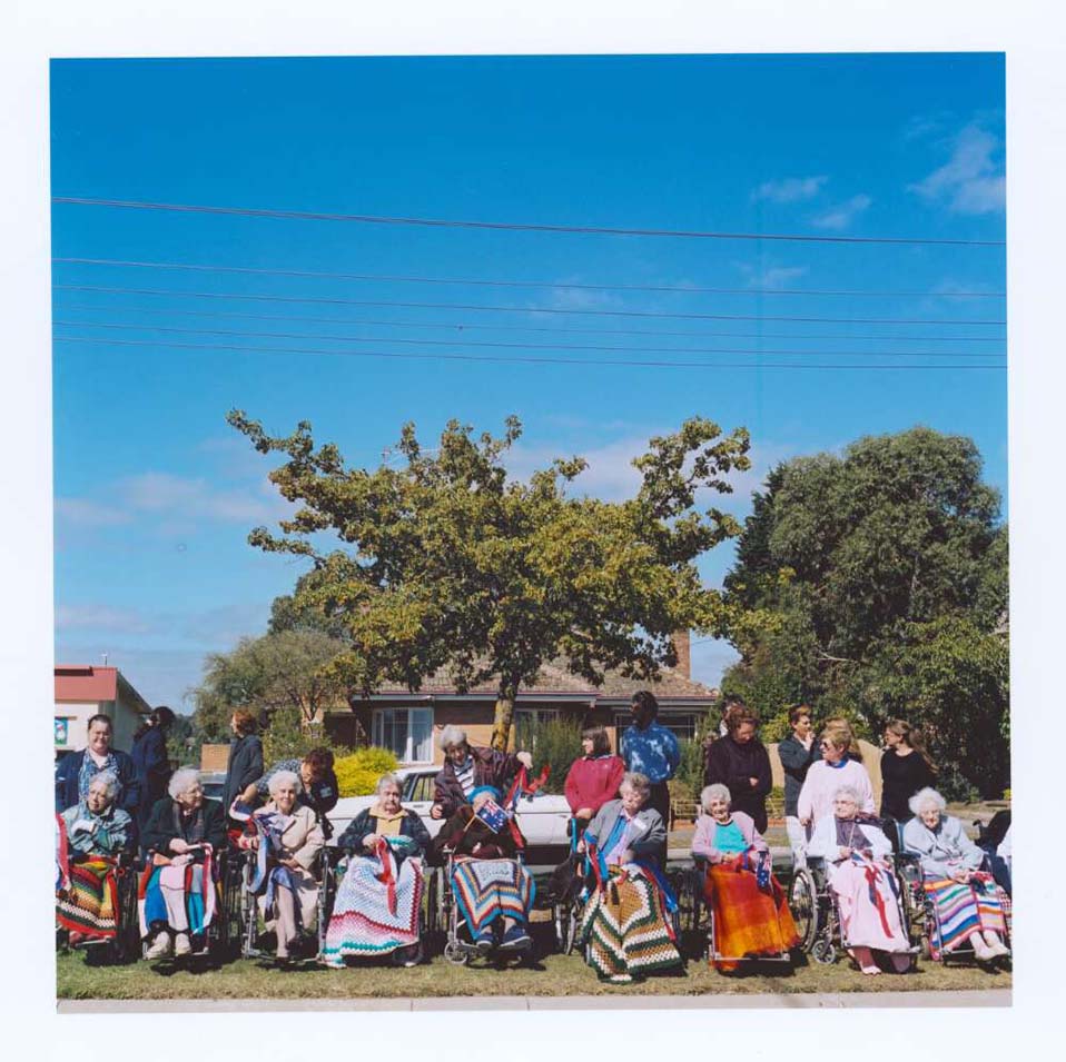 A bright sunny day with a group of elderly people sitting in a line in wheelchairs, all with knitted blankets over their knees.