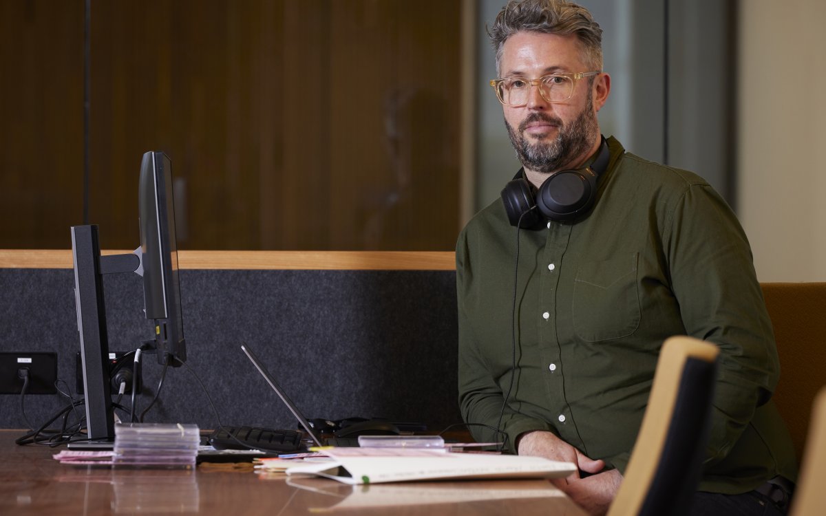 A man in glasses and a green shirt with headphones around his neck is sitting at a desk in front of a computer.