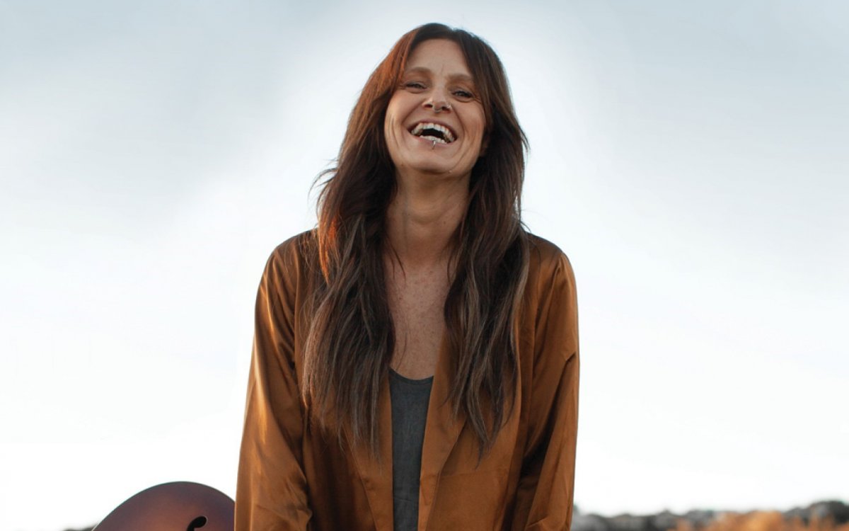 A woman with long brown hair, wearing a brown shirt and holding a guitar and smiling.