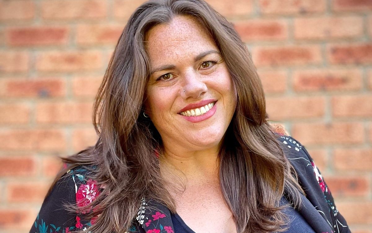 A headshot of a smiling woman with long, brown hair. She is wearing a colourful floral jack and is standing in front of a brick wall.