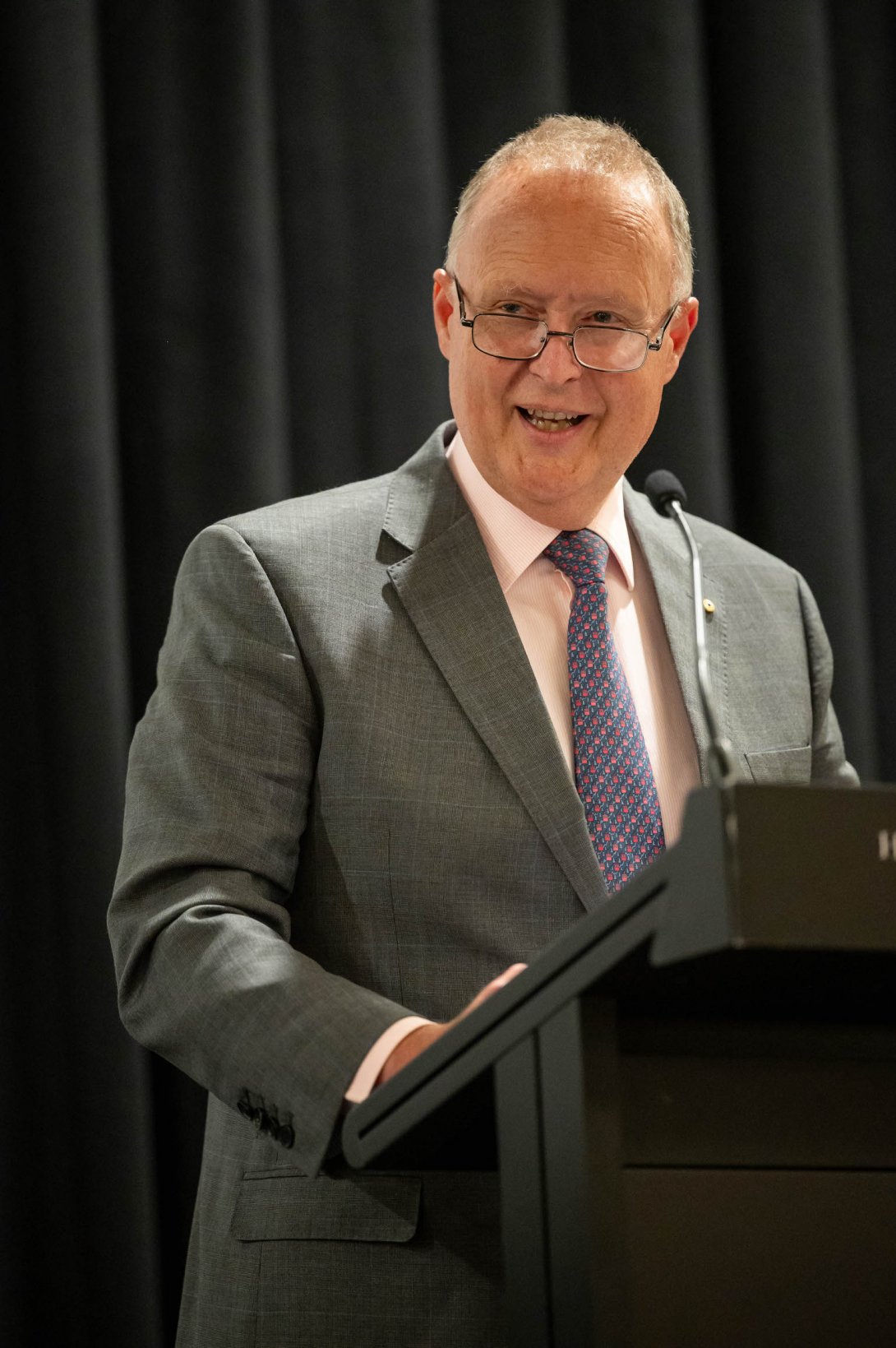 Rupert Myer AO, wearing a grey suit with a pink shirt and patterned tie, speaks at a podium with a microphone. He is smiling warmly and wearing glasses, addressing an audience against a backdrop of dark curtains.