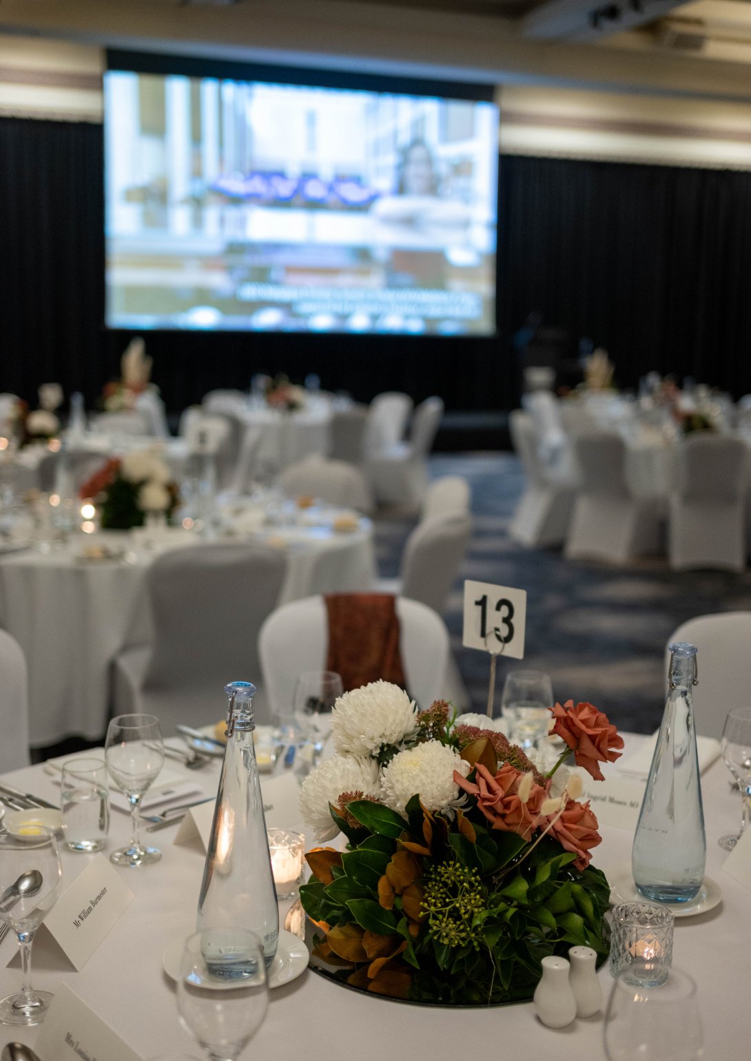 A beautifully set dinner table at the 2024 National Library of Australia Patron Dinner. The table features a floral centerpiece with white and orange flowers, leafy greenery, and small candles. The table is numbered '13' and surrounded by glassware, plates, and bottled water. In the background, more tables are set, and a large projector screen displays an image with blurred text