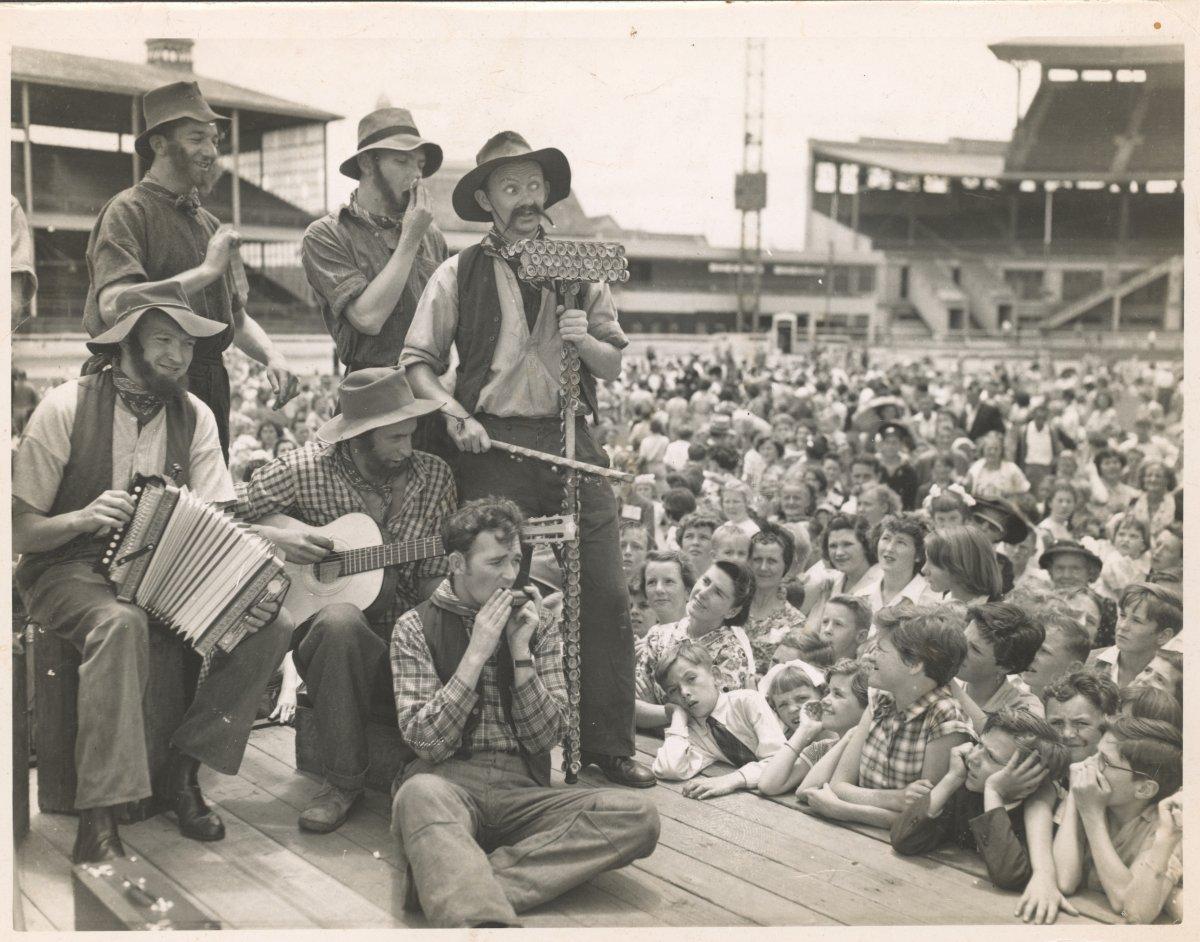 A group of performers play varied musical instruments atop a raised stage to a large crowd.