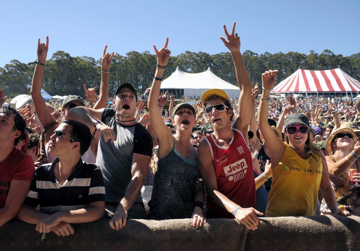 A crowd attend an outdoor music festival. They wear bright clothes, caps, sunglasses and have their hands raised in the air.