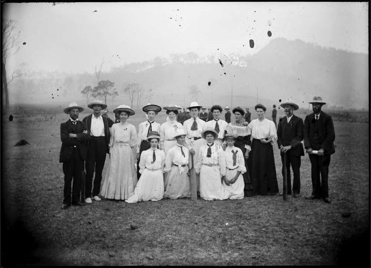 A black and white photo of 11 women standing in all white dresses flanked by 2 men on either side of them, dressed in black.