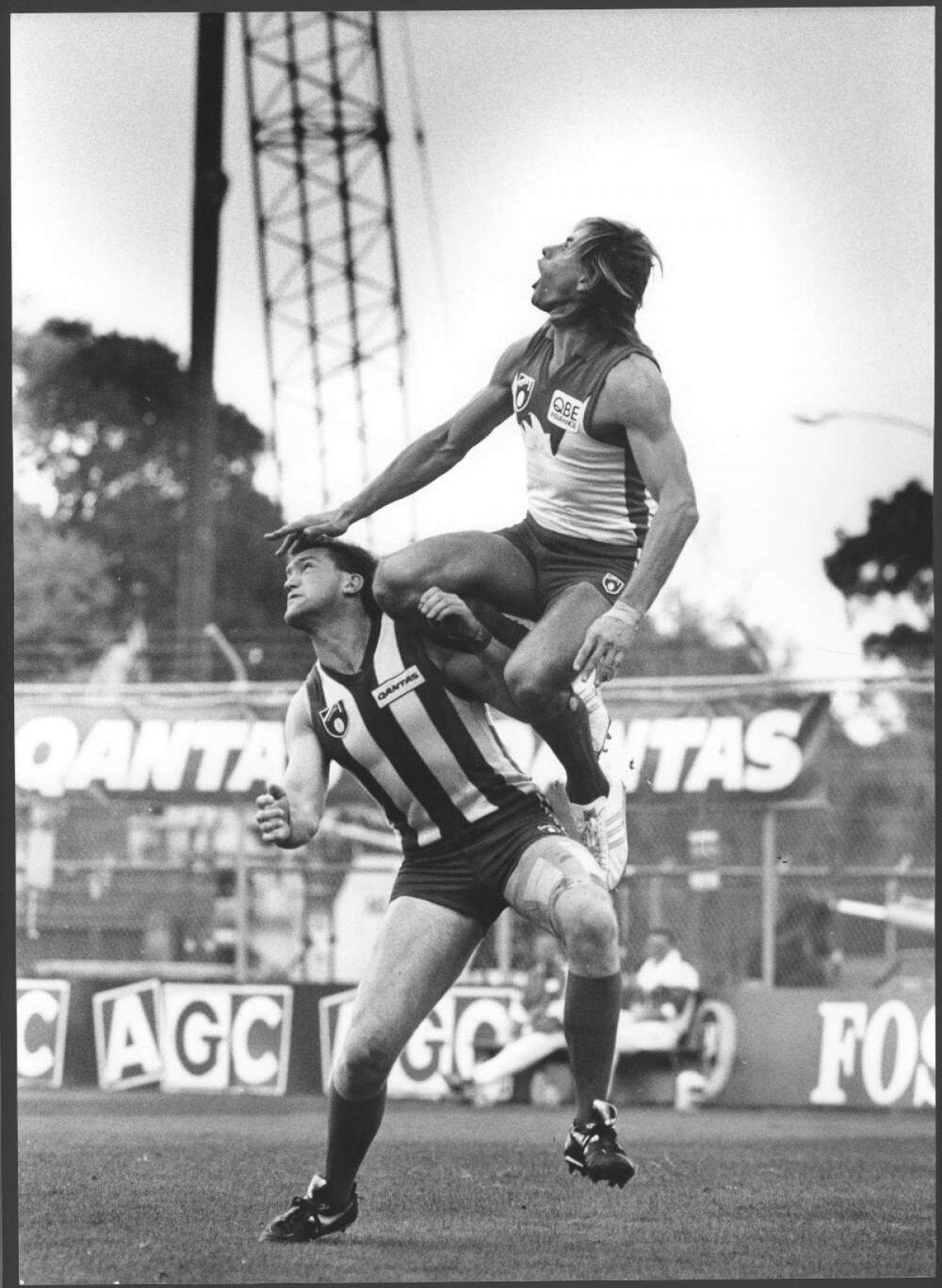 A black and white photo of Warwick Capper jumping high onto the shoulder of another player while looking up in the air.