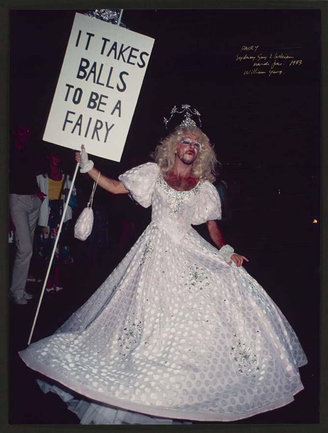 A participant at the 1993 Sydney Gay and Lesbian Mardi Gras wears a white ball gown with silver detailing and holds a sign reading 'It Takes Balls to be a Fairy.' The participant, with a blonde wig and makeup, strikes a playful pose, contributing to the lively atmosphere.