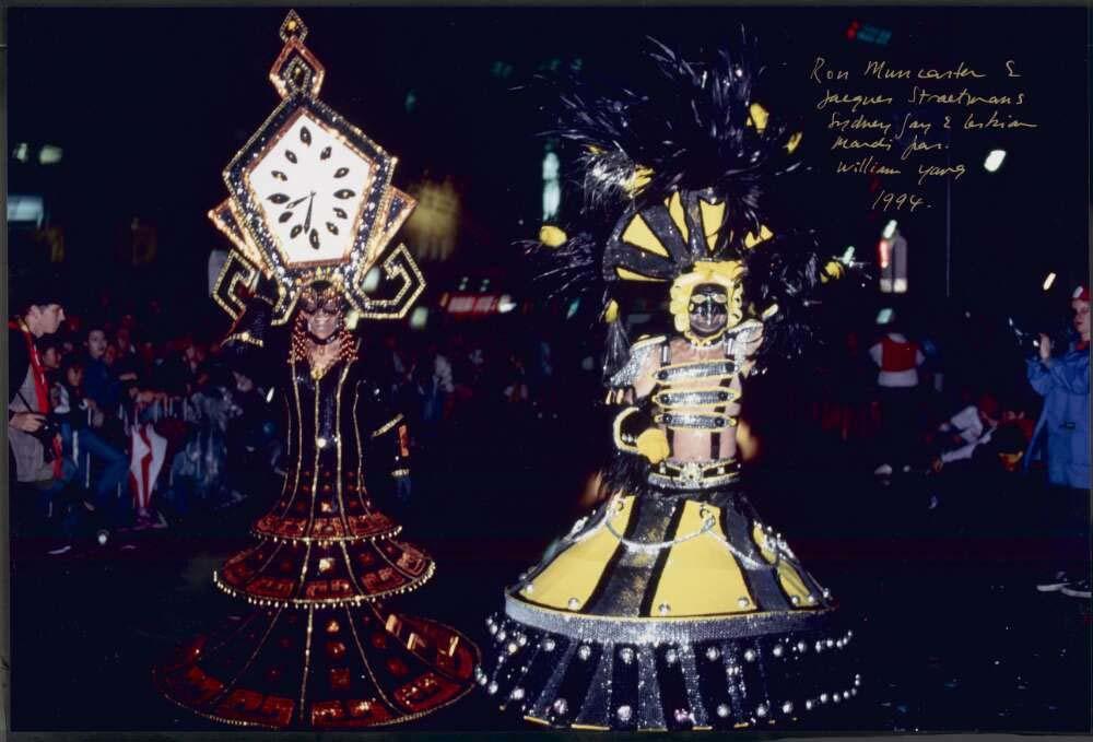 Two participants in elaborate costumes at the 1994 Sydney Gay and Lesbian Mardi Gras. The costume on the left resembles a large clock, with black and gold details. The right costume is yellow and black, with a feathered headdress and metallic elements.