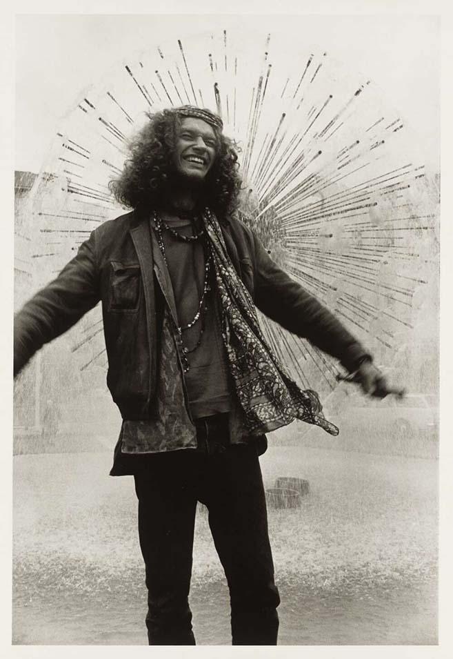 Black and white image of a smiling man with his arms spread wide standing in front of a fountain