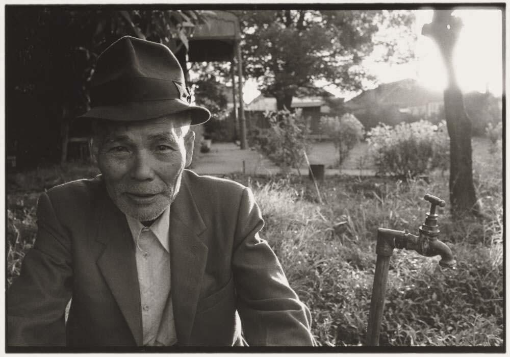 A black and white landscape of an older man wearing a hat and suit jacket sitting outside next to a water tap