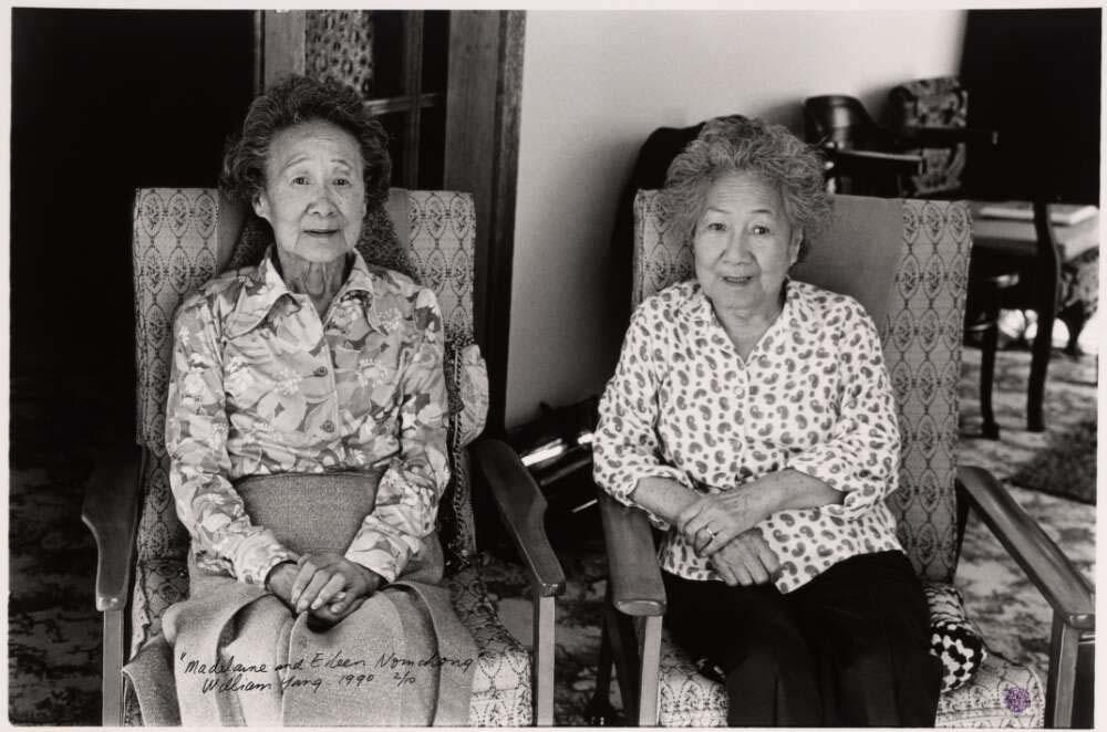 Two elderly women sitting in cushioned chairs looking at the camera with their hands folded in their laps.