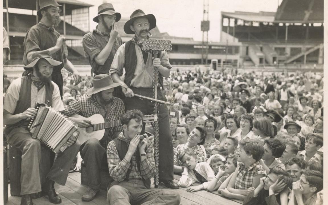 A group of performers play varied musical instruments atop a raised stage to a large crowd.