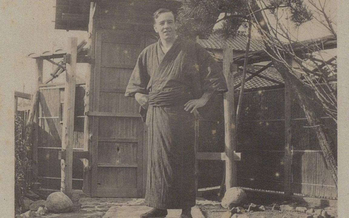 A sepia photograph of a man in traditional Japanese dress standing outside in front of a wooden dwelling structure.