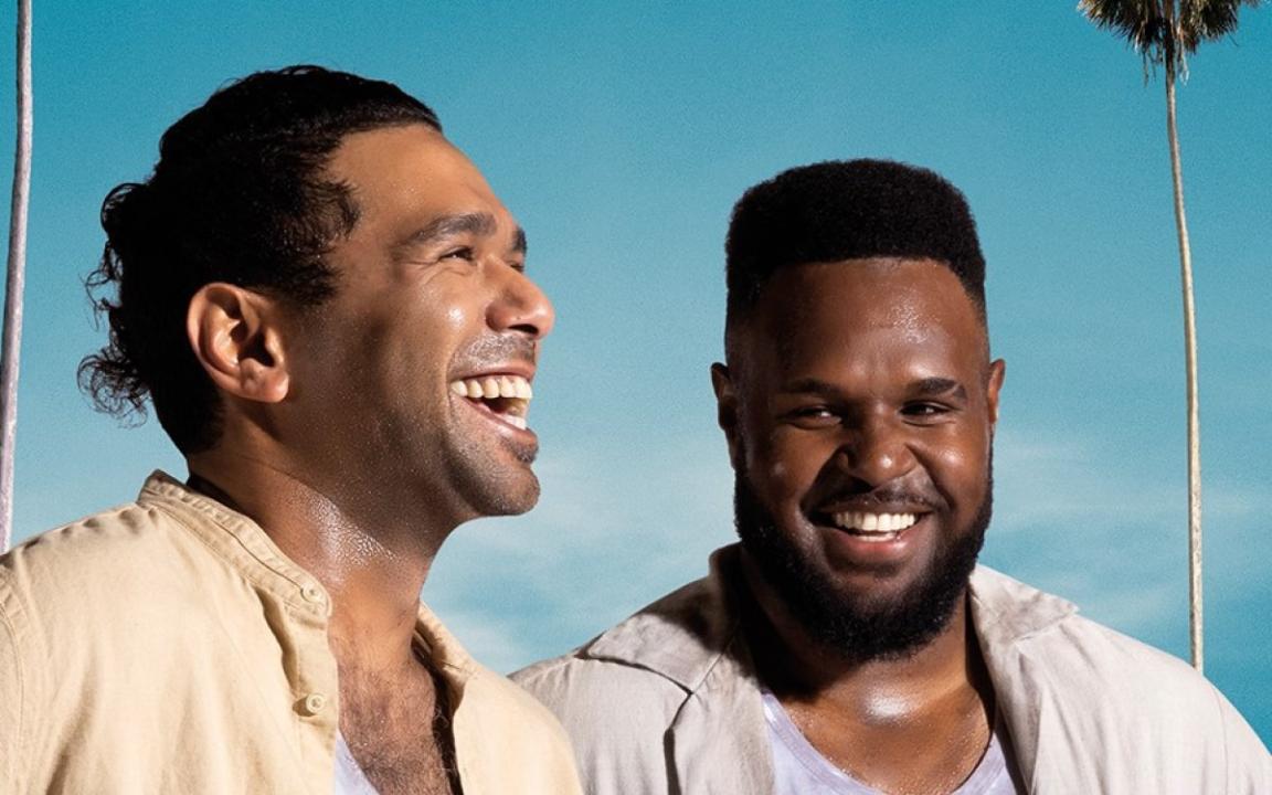 Two men smiling and laughing in front of a blue sky and palm tree.