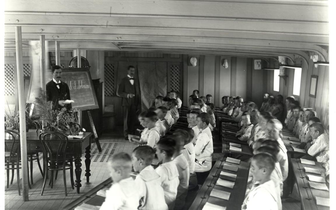 Classroom on a ship with many young boys sitting at long curved desks and two teachers standing at the front