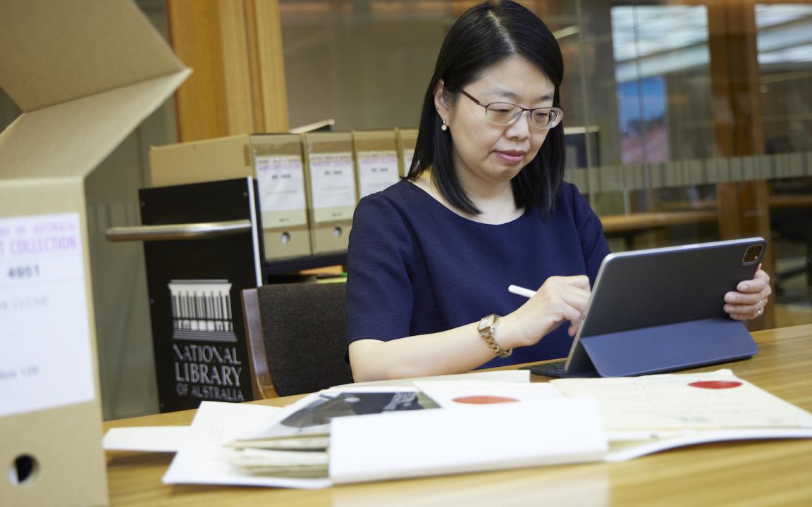 Woman with black hair and wearing a navy top using a laptop with a stylus at a desk covered in papers and a box