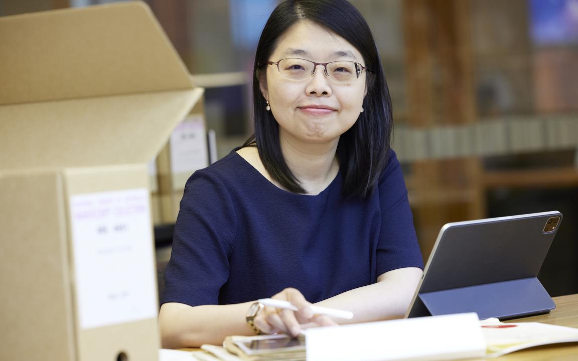 Woman with short dark hair wearing a navy short sleeve top smiling at the camera and sitting at a desk with a laptop, papers and a box