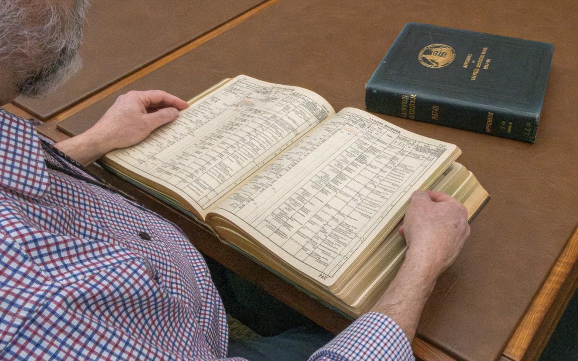 Man reading through a thick, leather-bound book of shipping registers