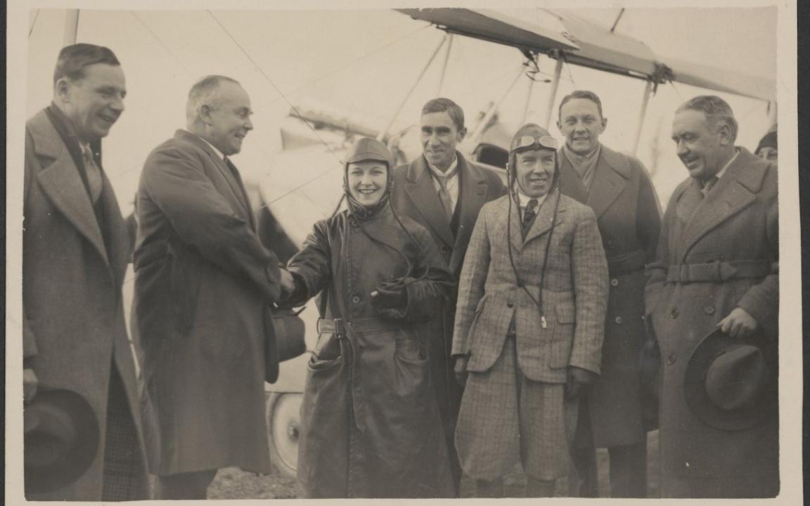 Female pilot in long coat smiling and shaking hands with a large man. Around them are 5 other smiling men. In the background, a wing of a plane can be seen