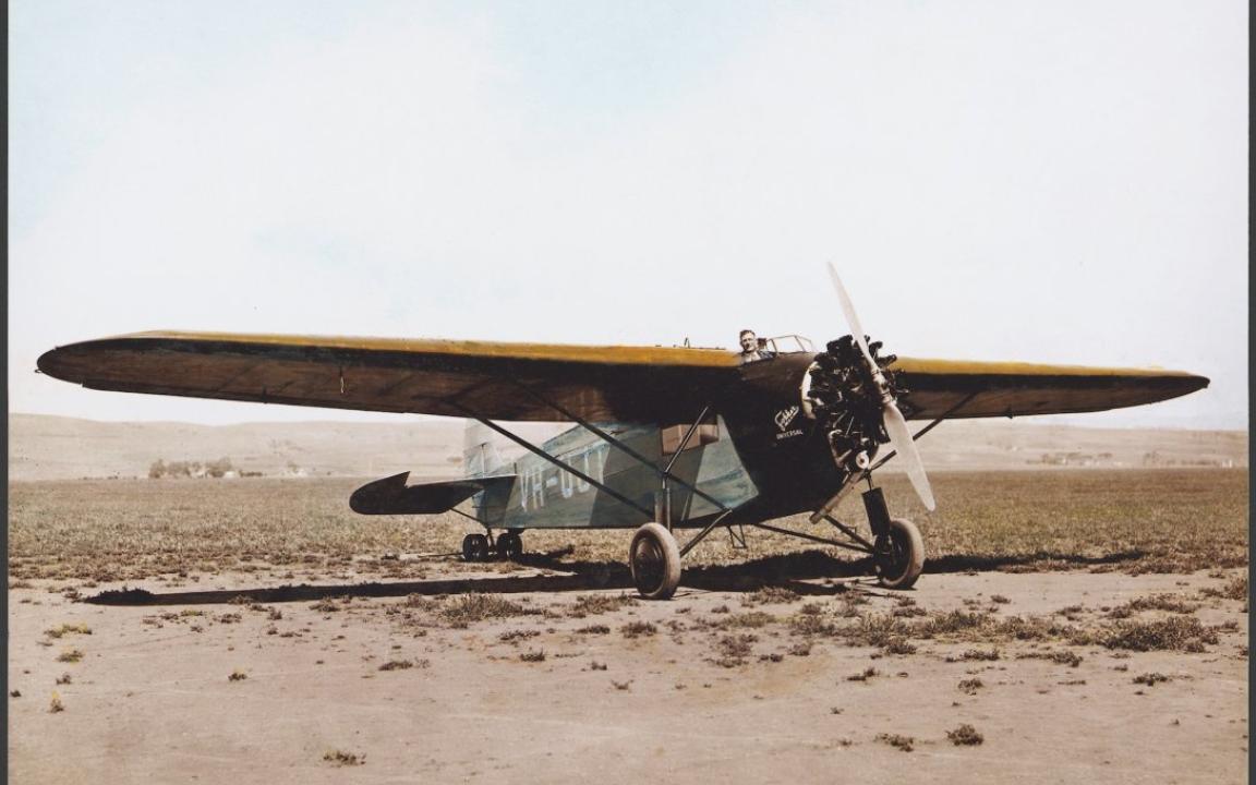 An old plane on the ground in a desert. A man is poking his head out from the cockpit and can be seen behind the wings of the plane