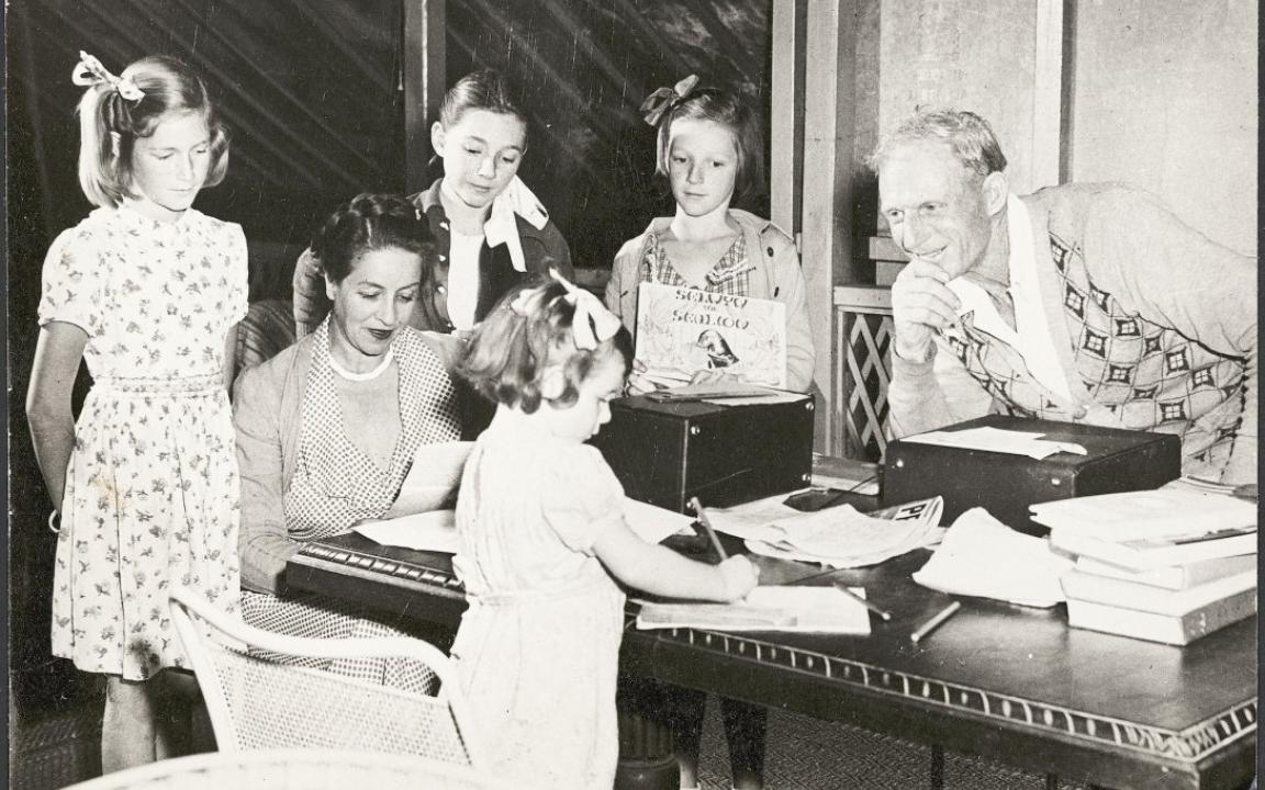 Family with mother, father and four young daughters sitting around a table with papers and boxes on it. The daughters al have bows in their hair and they all look content