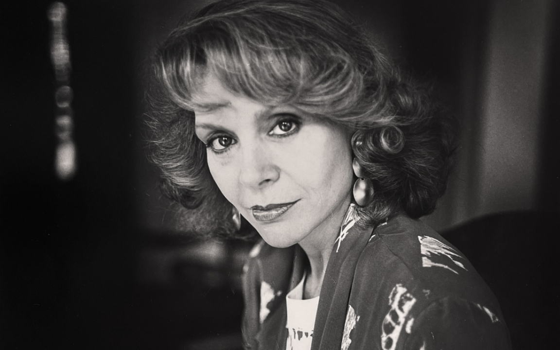 Black and white photograph of a woman with light, short curly hair looking directly into the camera with a small smile. She's wearing a dark blazer with light, abstract shapes on it and large round earrings