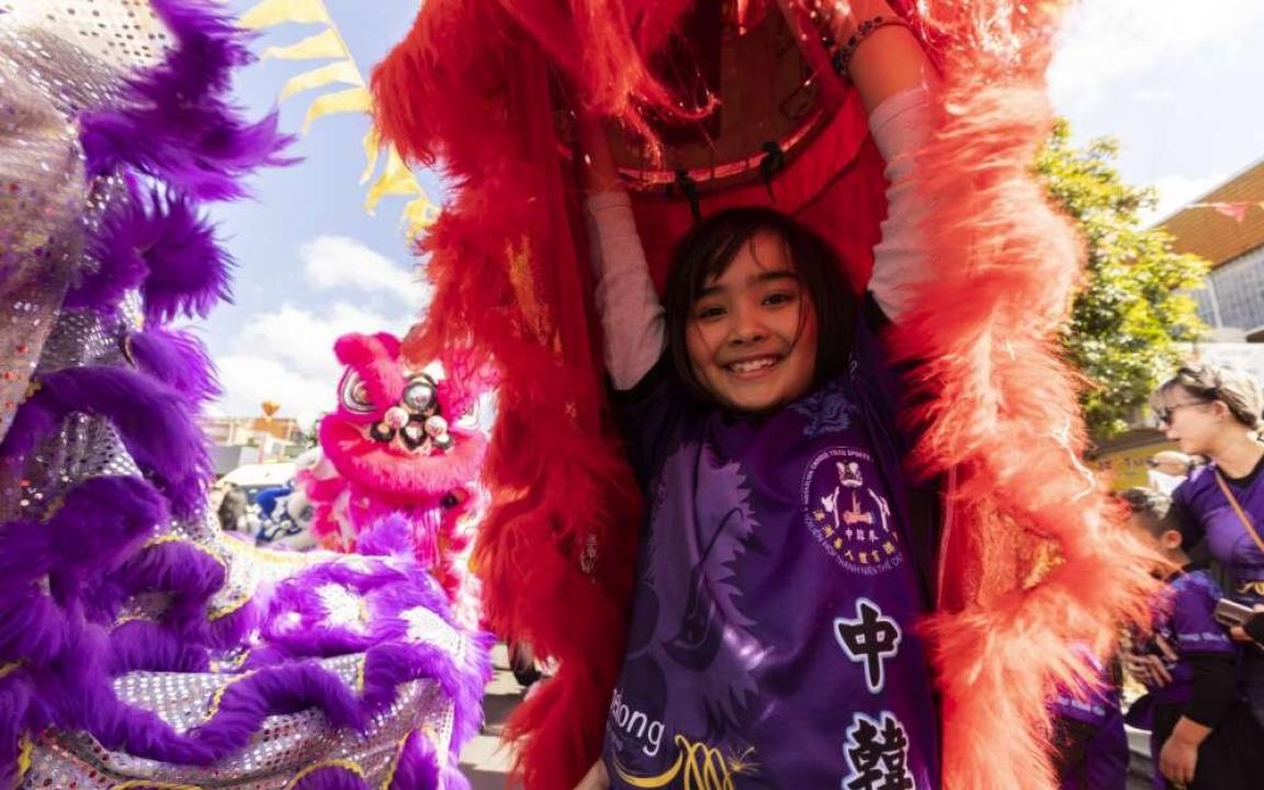 Young child in purple shirt smiles as she holds her lion costume for the Moon Festival above her head