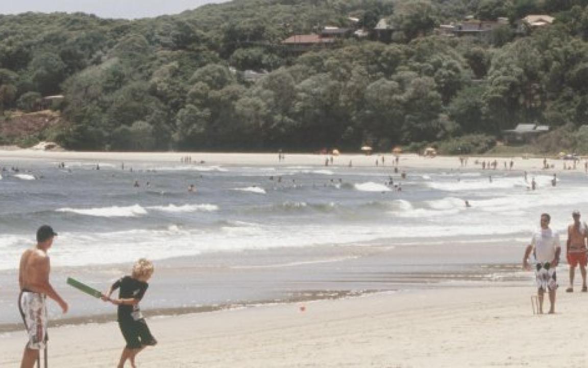 Group of men and a young boy playing cricket on the beach on a sunny, cloudless day.