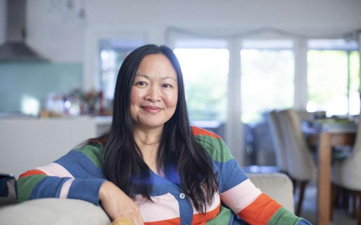 Woman of Chinese heritage sitting on a couch in a nice house, wearing a colourful striped jumper and smiling