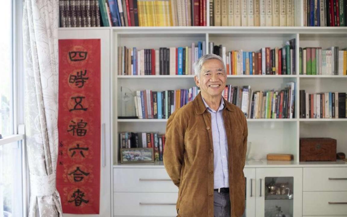 Older man of Chinese heritage in a brown jacket smiling and standing in front of a neat, colourful bookshelf