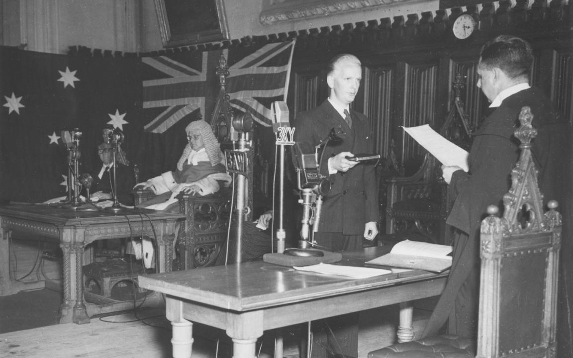 Man standing across a table from a man dressed in barrister's robes, with large United Kingdom and Australian flags hung on the wall behind him