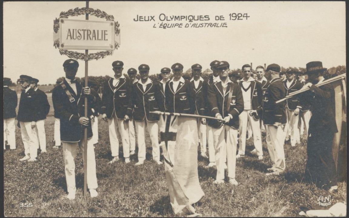 Group of men in white pants and dark blazers standing in a field. One of them holds a previous version of the Australian flag and another a sign that Reads 'Australie'