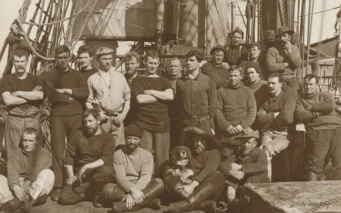 Sepia toned photo of a chips crew made up of about 20 men and a dog, on the deck of their ship 'Terra Nova'