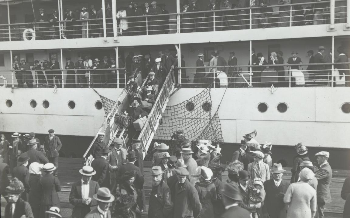 Passengers with luggage crossing a bridge between a large boat and a dock, with people looking on from the upper level of the boat and lots of people waiting around the dock