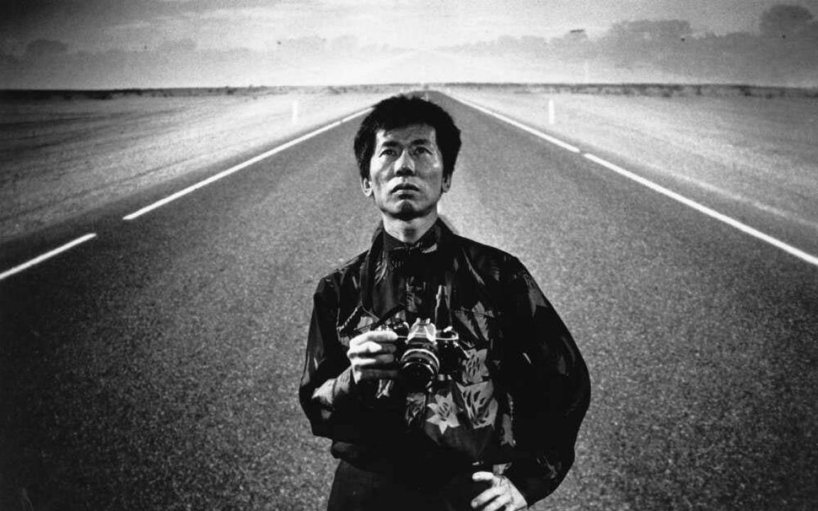 A black-and-white portrait of William Yang standing alone on an empty road, holding a camera. The road stretches behind Yang as he gazes straight into the camera in a solemn pose.