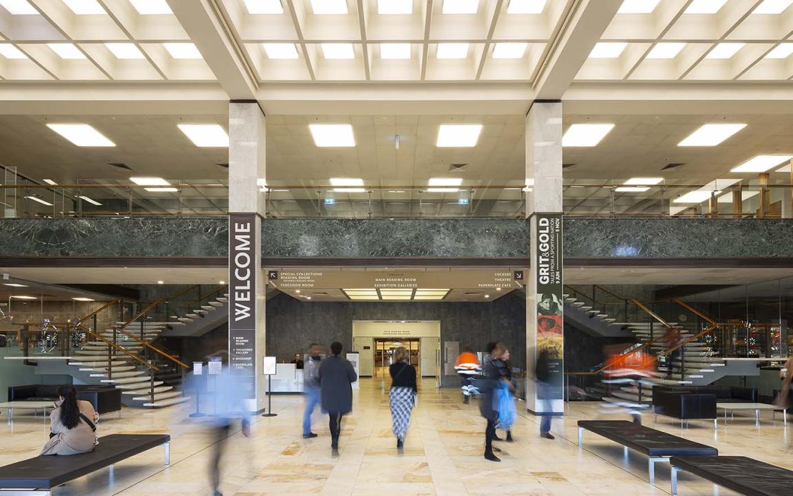 Large two-story room with cream-coloured marble floors, columns and stairs; black leather couches; lots of light from the ceiling and a balcony from the second floor looking over the room. On the columns are signs with directions and the word 'Welcome'. People are moving around the room.