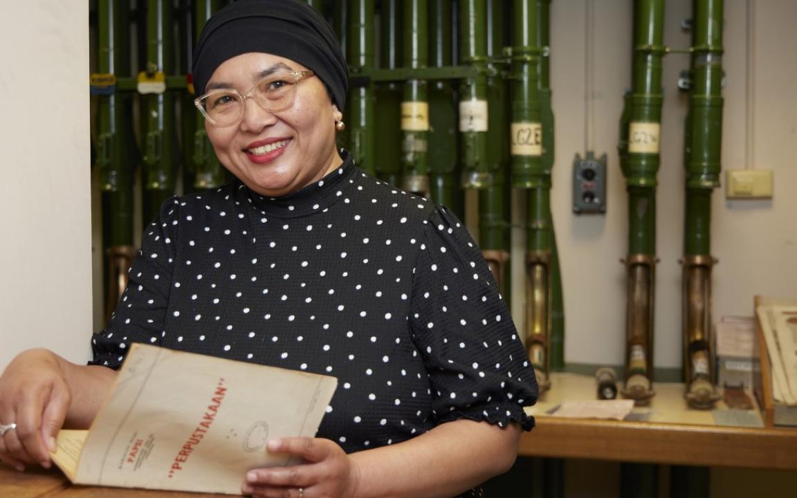 Woman wearing a dot print shirt, glasses and a black head scarf holding an open booklet and smiling