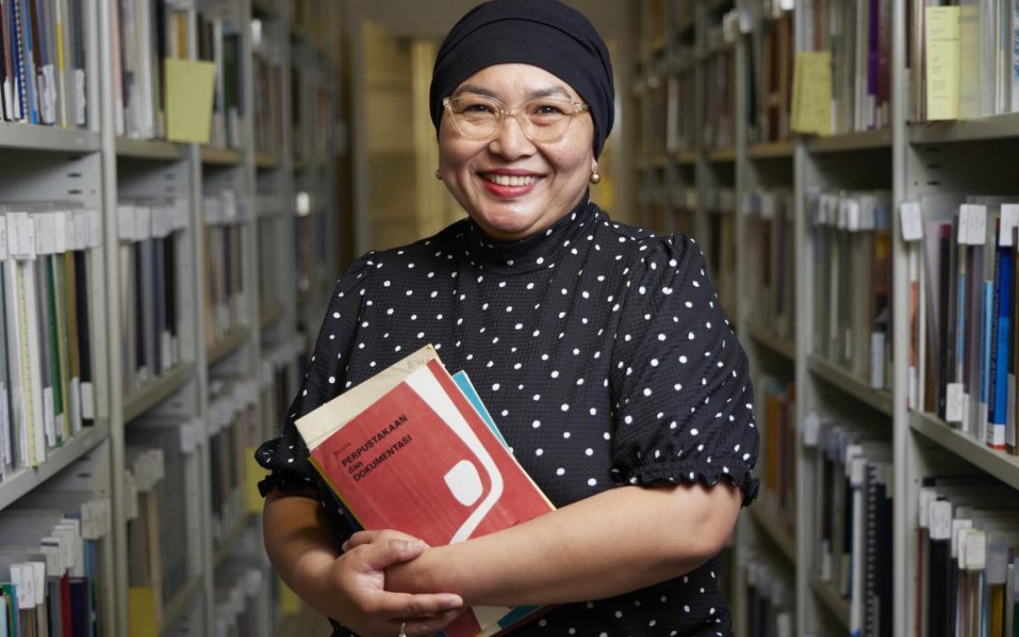 Woman wearing a dot print shirt, glasses and a black head scarf holding some booklets in an aisle of shelves