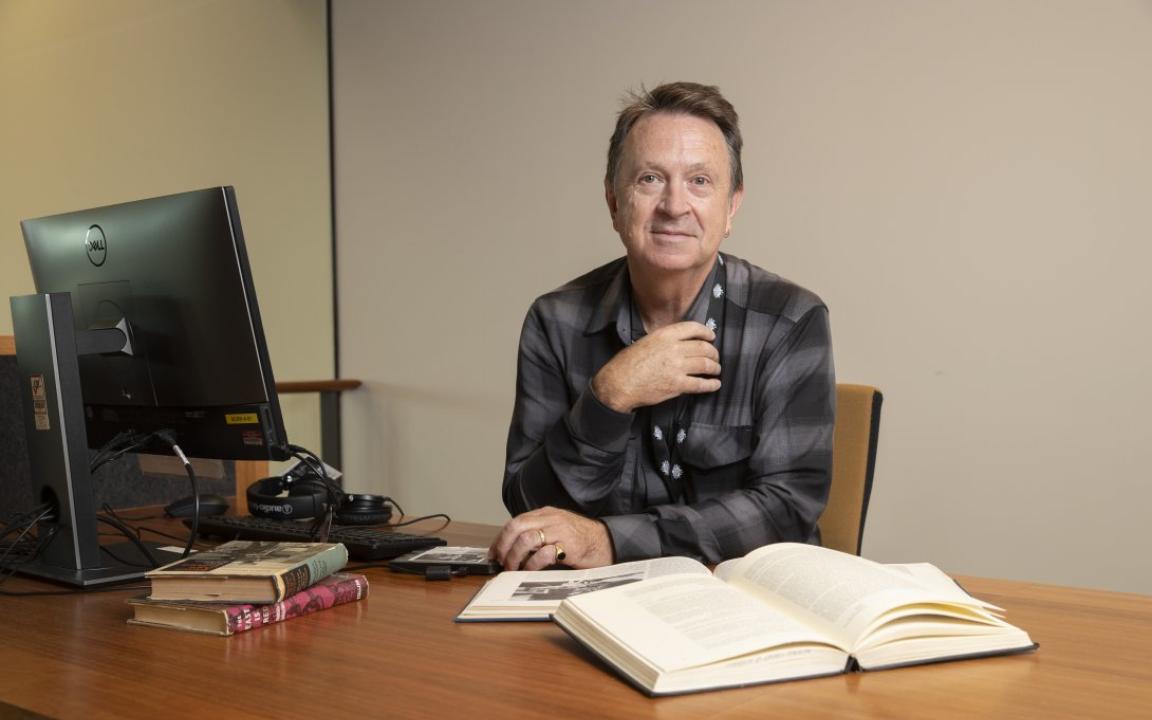 A man wearing a black and grey checkered shirt sitting at a desk with a computer and several books resting on it. He is leaning on the desk, facing forward and giving the camera a small smile