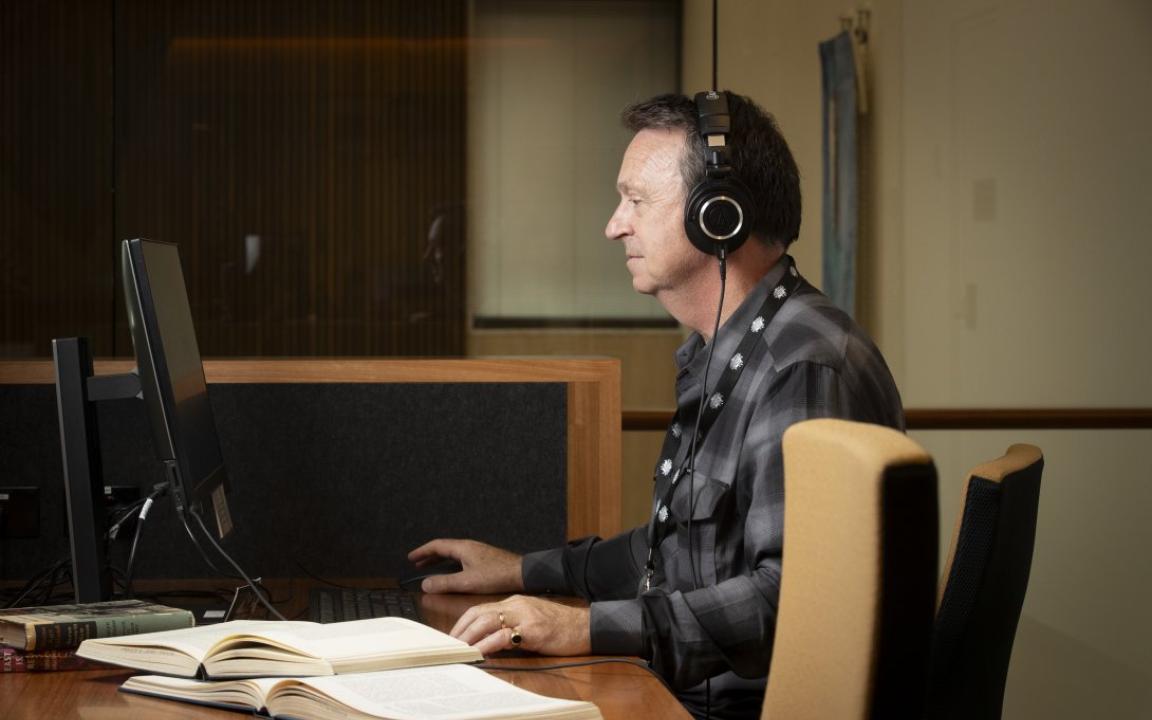 Man in a grey and black checkered shirt and headphones sitting at a desk, using a computer. To his left are two open books