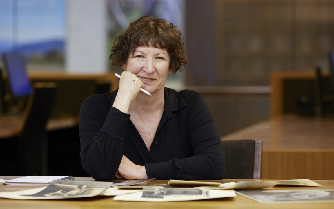 Woman with short, curly brown hair wearing a black top sitting at a table covered in old photos and manuscripts, smiling