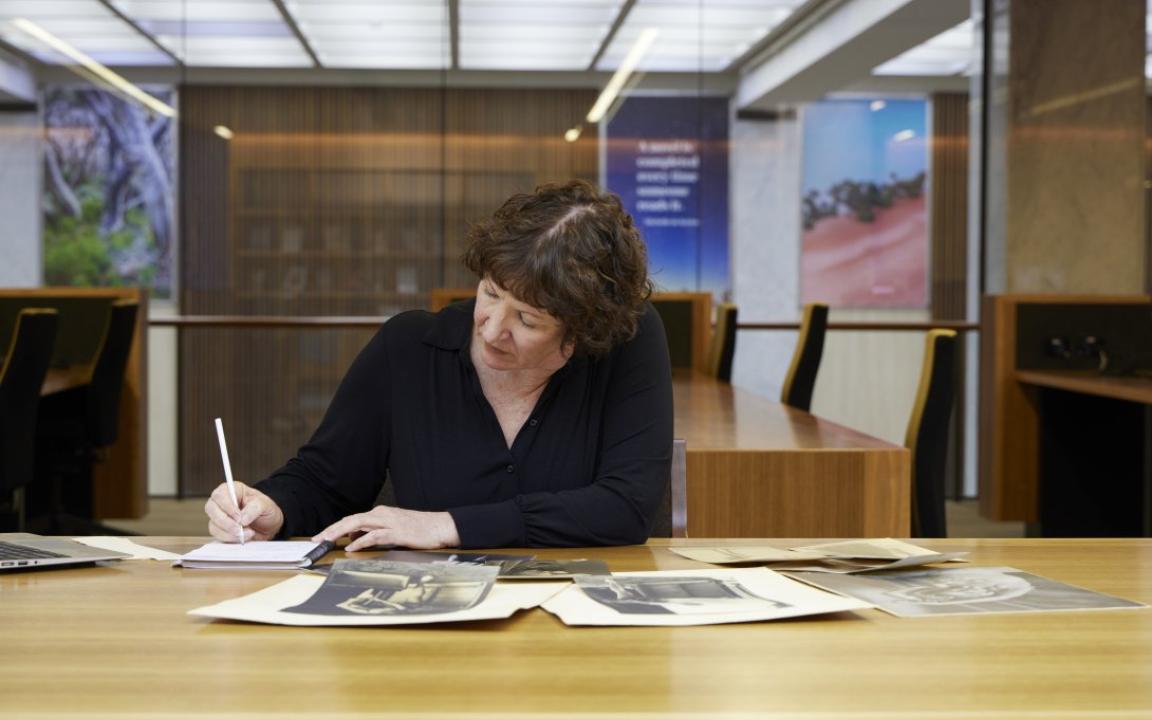 Woman with short, curly brown hair wearing a black top sitting at a table covered in old photos and manuscripts, writing in a notebook