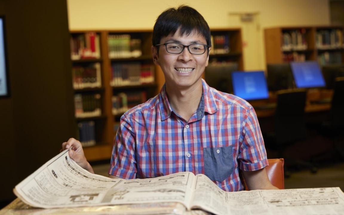 Man with short black hair and black glasses in a short-sleeved checkered shirt smiling and sitting at a desk with a Chinese language newspaper on it